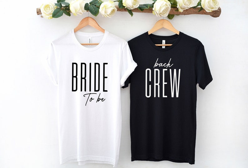 May include: Two white t-shirts with black text. One shirt says "Bride to be" and the other says "Bach Crew".