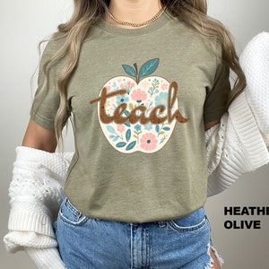 Op de afbeelding: Een groen T-shirt met een bloemachtig appelontwerp en het woord "teach" in bruine letters. Het shirt is helio-olijfkleurig.
