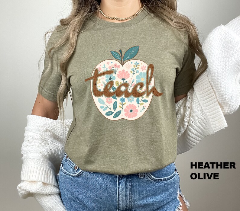 Op de afbeelding: Een groen T-shirt met een bloemachtig appelontwerp en het woord "teach" in bruine letters. Het shirt is helio-olijfkleurig.