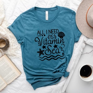 May include: A teal blue t-shirt with the text "All I need is Vitamin Sea" in black. The text is surrounded by a seashell, a starfish, and wavy lines.