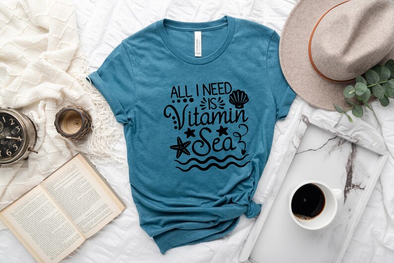 May include: A teal blue t-shirt with the text "All I need is Vitamin Sea" in black. The text is surrounded by a seashell, a starfish, and wavy lines.