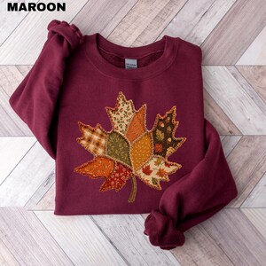 Pode incluir: Uma camisola de manga comprida cor de vinho com uma grande aplica&ccedil;&atilde;o central de folha de &aacute;cer. A folha &eacute; constru&iacute;da a partir de v&aacute;rios retalhos de tecido em tons de laranja, castanho, verde e creme, com padr&otilde;es distintos e bordos cosidos.