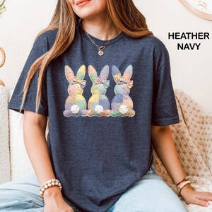 May include: Heather navy blue t-shirt with a design of three patchwork bunny rabbits, each with a bow and colourful patterns. The text "HEATHER NAVY" is printed on the right. A casual, comfortable style.
