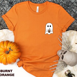 Puede incluir: Camiseta naranja quemada con un fantasma blanco sosteniendo una taza de caf&eacute;. El fantasma tiene una sonrisa amigable.