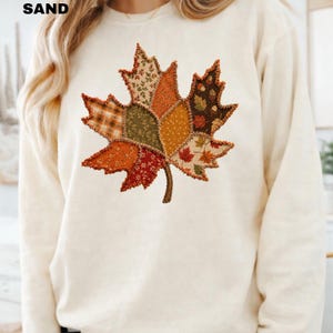 Pode incluir: Uma camisola de gola redonda cor de areia com um grande gr&aacute;fico central de folha de &aacute;cer composto por v&aacute;rios padr&otilde;es de tecido patchwork em tons de laranja, castanho, verde e creme, com costuras vis&iacute;veis ao longo das bordas da folha.