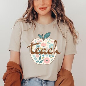 Op de afbeelding: Een beige T-shirt met een bloemachtig appelontwerp en het woord "teach" in bruine tekst.