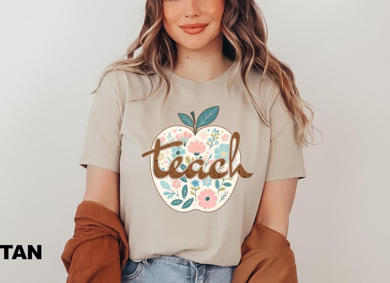Op de afbeelding: Een beige T-shirt met een bloemachtig appelontwerp en het woord "teach" in bruine tekst.