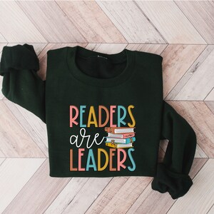May include: A dark green sweatshirt with the text "Readers are Leaders" in colorful letters. A stack of colorful books is pictured next to the text.
