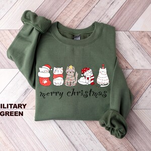 May include: A green sweatshirt with a festive design featuring five cats wearing Christmas hats and decorations. The text "merry christmas" is printed below the cats.