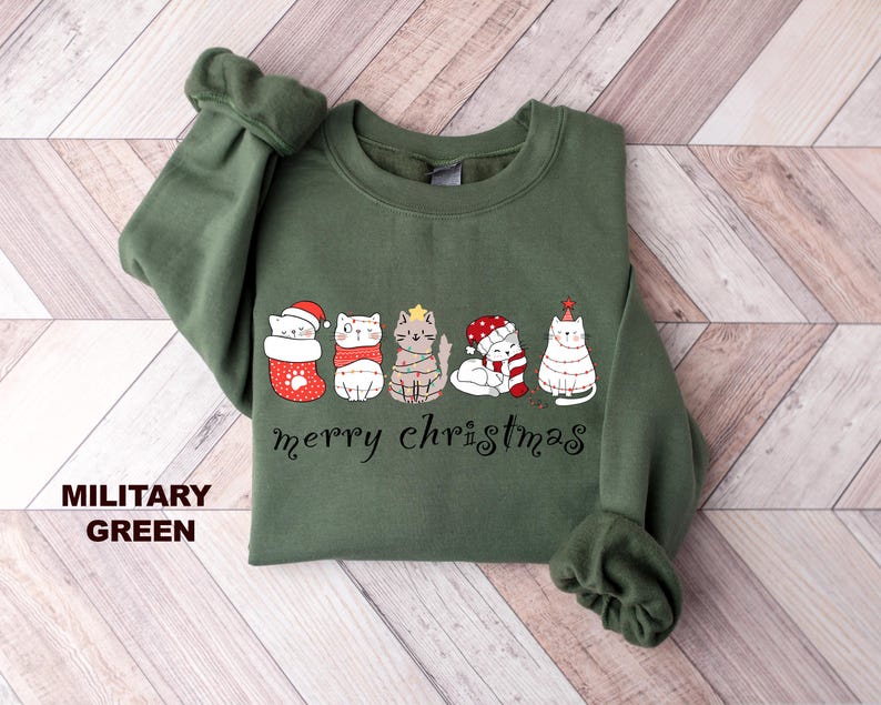 May include: A green sweatshirt with a festive design featuring five cats wearing Christmas hats and decorations. The text "merry christmas" is printed below the cats.