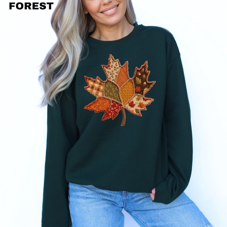 Pode incluir: Uma camisola de gola redonda verde-floresta com um grande gr&aacute;fico central de uma folha de &aacute;cer. A folha &eacute; composta por v&aacute;rios segmentos de tecido estilo patchwork em tons de laranja, castanho e dourado, com um rebordo cosido.