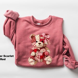 May include: Heather scarlet red sweatshirt with a patchwork teddy bear design. The bear has a heart that says "Love" and the words "Hugs" and "Kisses" on its paws. The bear is made of various patterned fabrics.