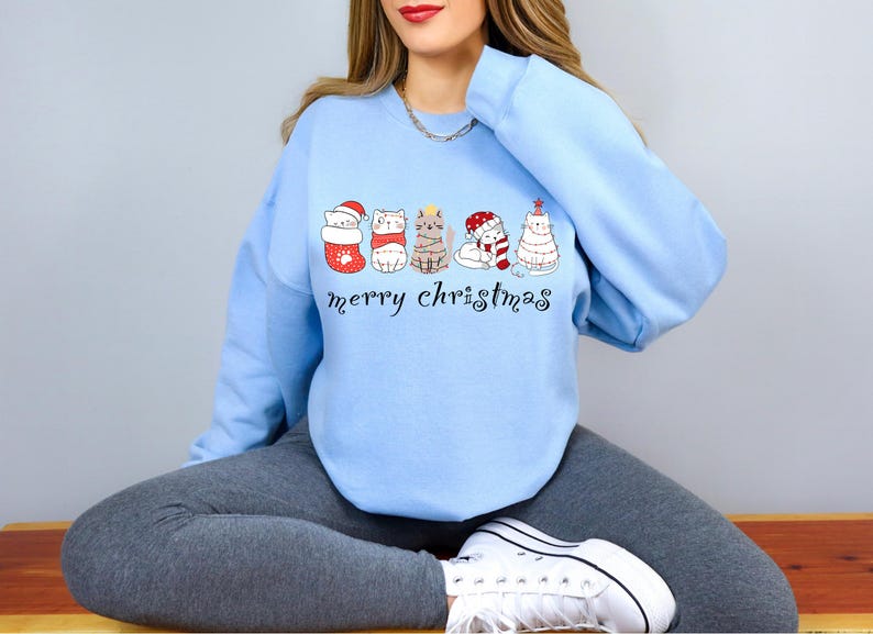 May include: A light blue sweatshirt with a Christmas-themed design featuring a line of cats wearing Santa hats, stockings, and a Christmas tree. The text "merry christmas" is printed below the cats.