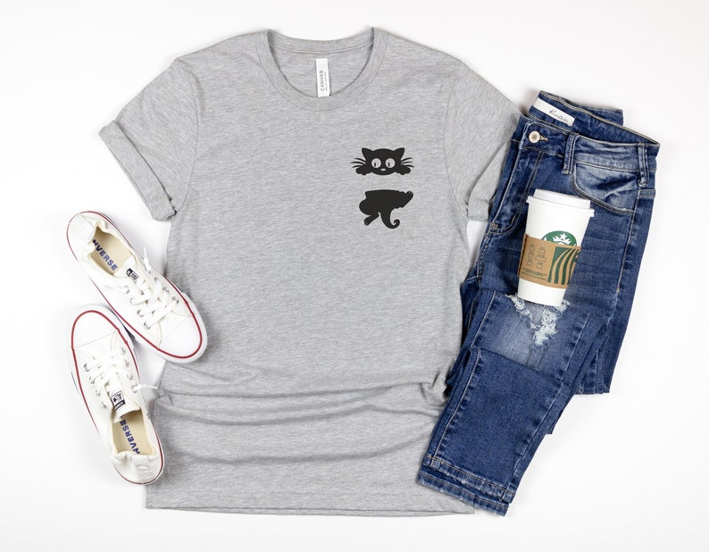 May include: A gray t-shirt with a black cat silhouette graphic on the front. The cat is peeking out from behind a wall. The shirt is paired with blue jeans, white sneakers, and a coffee cup.