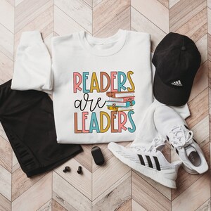 May include: White sweatshirt with the text "Readers are Leaders" in colorful letters. A stack of books is pictured above the word "Leaders". The sweatshirt is laid out on a wood floor with black leggings, a black baseball cap, and white sneakers.