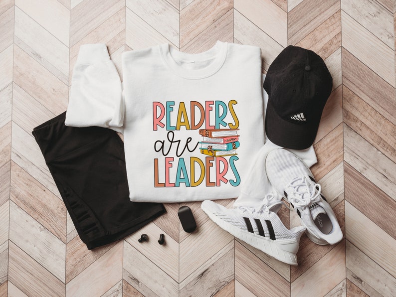 May include: White sweatshirt with the text "Readers are Leaders" in colorful letters. A stack of books is pictured above the word "Leaders". The sweatshirt is laid out on a wood floor with black leggings, a black baseball cap, and white sneakers.