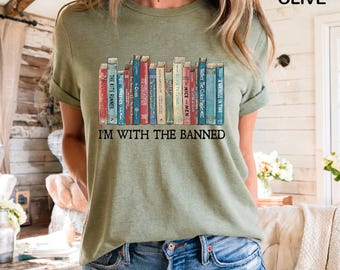 May include: Olive green t-shirt featuring a graphic of a row of colorful books with titles like "The Kite Runner" and "A Wrinkle in Time." The text "I'M WITH THE BANNED" is below the books. The shirt is made of soft material.