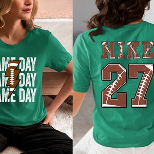 May include: Two green t-shirts with a football theme. One shirt says "Game Day" three times with a football and lightning bolt graphic. The other shirt has the name "Mike" and the number "27" with a football graphic.