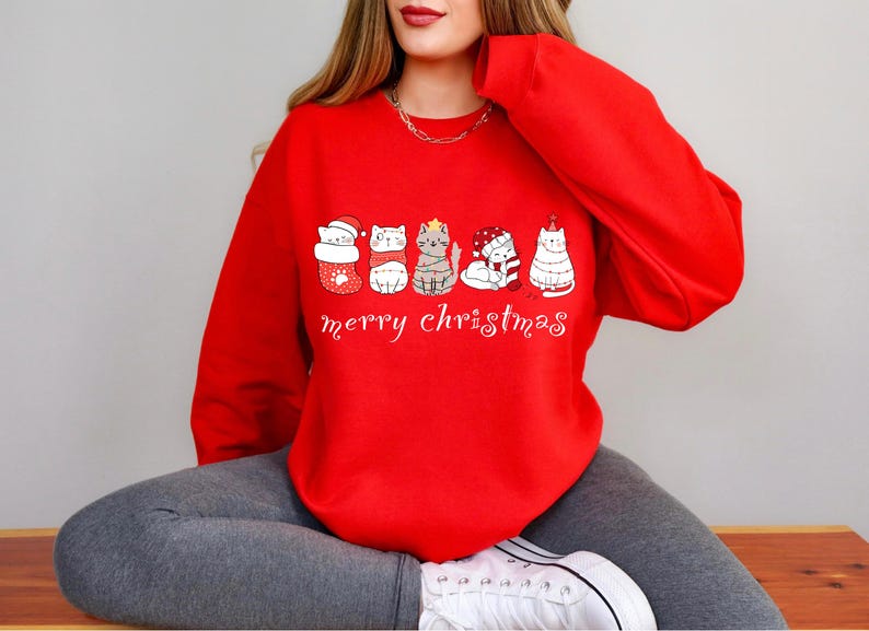 May include: Red Christmas sweatshirt with a white graphic of cats wearing Santa hats and Christmas tree costumes. The text "merry christmas" is printed below the cats.