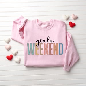 Peut inclure: Sweat-shirt rose avec le texte "girls weekend" en couleurs arc-en-ciel.