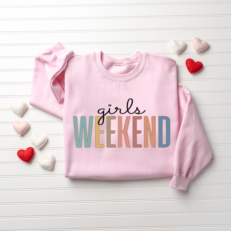 Peut inclure: Sweat-shirt rose avec le texte "girls weekend" en couleurs arc-en-ciel.