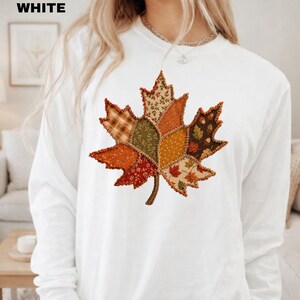 Pode incluir: Uma camisa branca de manga comprida com um grande gr&aacute;fico central de uma folha de bordo composta por v&aacute;rios padr&otilde;es de tecido estilo patchwork em tons de laranja, castanho, verde e amarelo, com bordas costuradas vis&iacute;veis.