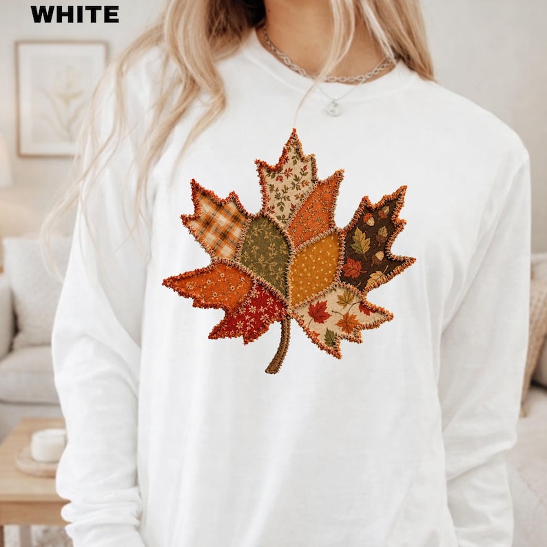 Pode incluir: Uma camisa branca de manga comprida com um grande gr&aacute;fico central de uma folha de bordo composta por v&aacute;rios padr&otilde;es de tecido estilo patchwork em tons de laranja, castanho, verde e amarelo, com bordas costuradas vis&iacute;veis.