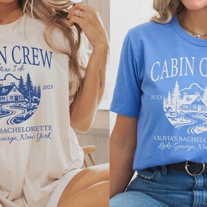 May include: Two short-sleeve t-shirts, one cream with blue text and one blue with white text. Both feature a circular graphic of a cabin in the woods with the text CABIN CREW, before I do, JULY 2025, OLIVIA'S BACHELORETTE, and Lake George, New York.