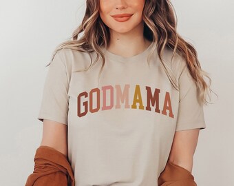 May include: A beige t-shirt with the text "GODMAMA" in a rainbow colour gradient.