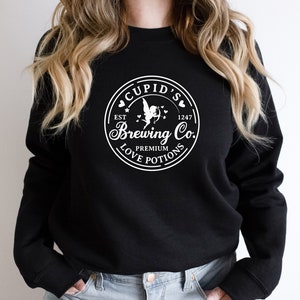 May include: Black sweatshirt with a white graphic of a circle with the text "Cupid's Brewing Co." inside. The graphic also includes a cupid with wings and arrows, hearts, and the text "Est 1247" and "Premium Love Potions".