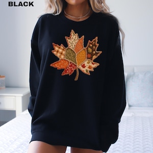 Pode incluir: Uma camisola preta de manga comprida com um grande gr&aacute;fico central de uma folha de &aacute;cer. A folha &eacute; composta por v&aacute;rios segmentos de tecido estilo patchwork em cores de outono, incluindo laranja, castanho, verde e creme, com padr&otilde;es florais.