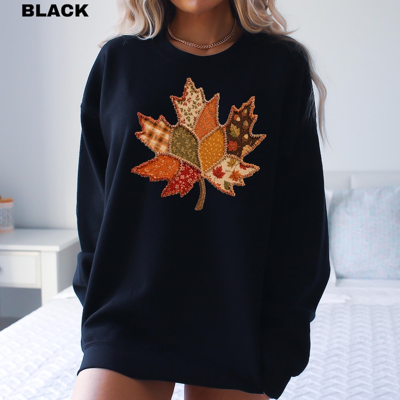 Pode incluir: Uma camisola preta de manga comprida com um grande gr&aacute;fico central de uma folha de &aacute;cer. A folha &eacute; composta por v&aacute;rios segmentos de tecido estilo patchwork em cores de outono, incluindo laranja, castanho, verde e creme, com padr&otilde;es florais.
