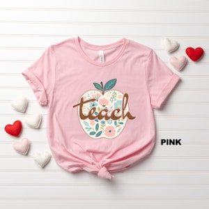 Op de afbeelding: Roze T-shirt met een bloemachtig appelontwerp en het woord "teach" in bruine tekst.