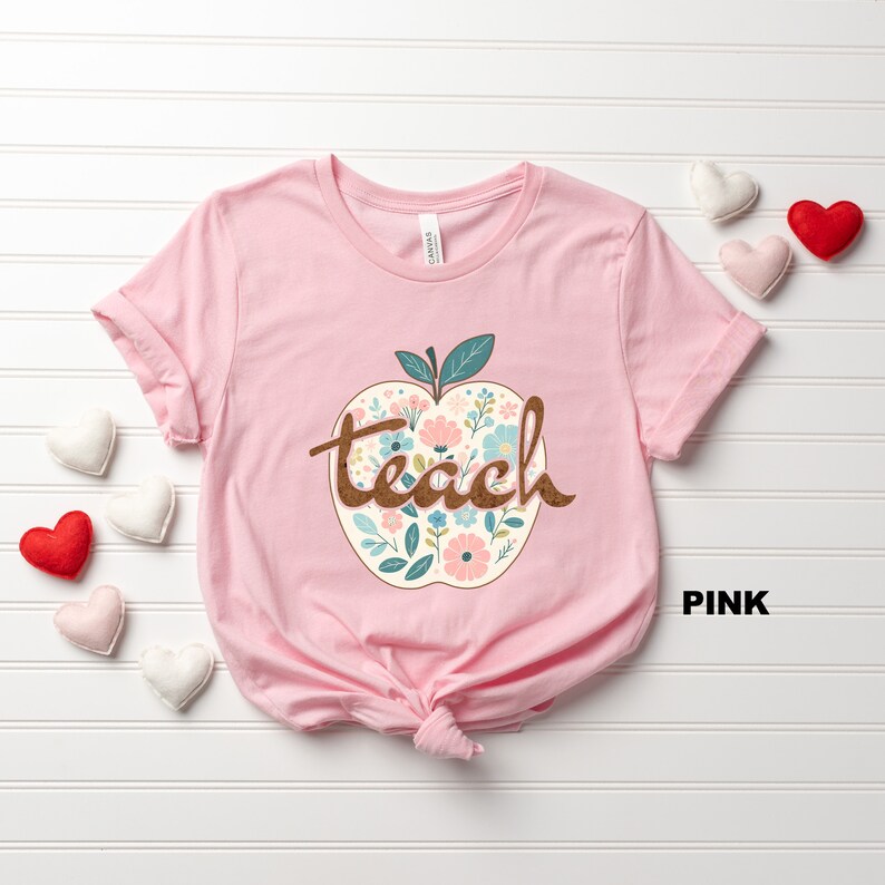 Op de afbeelding: Roze T-shirt met een bloemachtig appelontwerp en het woord "teach" in bruine tekst.