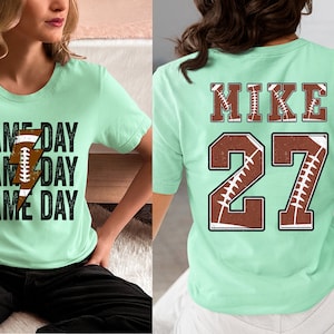 May include: Two mint green t-shirts with a football theme. One shirt has the text "GAME DAY" repeated three times with a football graphic. The other shirt has the text "MIKE 27" with football graphics.