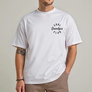 May include: White t-shirt with the text "Cool Grandpas Club" in a black, circular design. The shirt has short sleeves and a crew neck. The model is wearing brown pants and a bracelet.