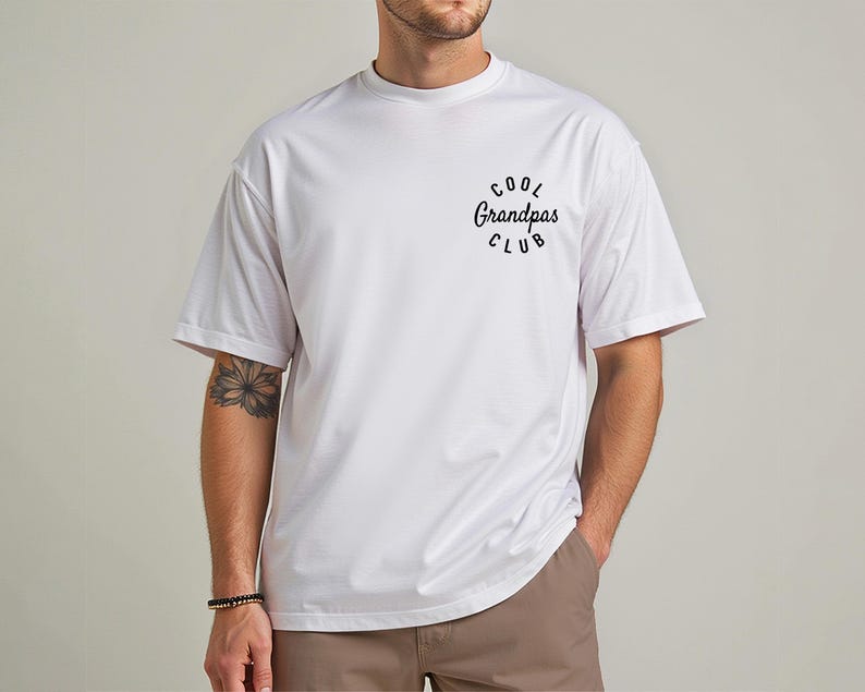 May include: White t-shirt with the text "Cool Grandpas Club" in a black, circular design. The shirt has short sleeves and a crew neck. The model is wearing brown pants and a bracelet.