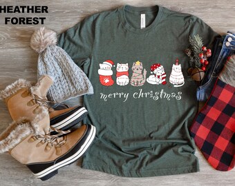 May include: A heather forest green t-shirt with a "merry christmas" graphic featuring six cartoon cats in festive attire. The image includes a gray knit hat, tan winter boots, and a red and black plaid scarf.