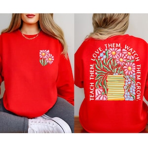 May include: A red sweatshirt with a floral design on the front and back. The front features two embroidered daisies. The back has a floral arrangement with the text "Teach Them, Love Them, Watch Them Grow."