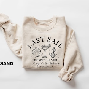 May include: Sand-colored sweatshirt with the text "LAST SAIL" above a seashell, cocktail, and anchor graphic. Below the graphic, the text reads "BEFORE THE VEIL Megan's Bachelorette SAN DIEGO, CA". The sweatshirt's sleeves are folded.