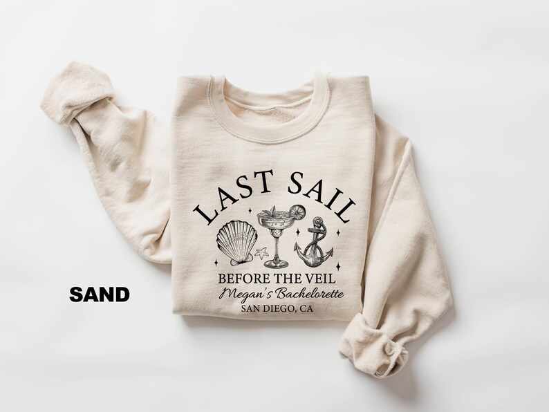 May include: Sand-colored sweatshirt with the text "LAST SAIL" above a seashell, cocktail, and anchor graphic. Below the graphic, the text reads "BEFORE THE VEIL Megan's Bachelorette SAN DIEGO, CA". The sweatshirt's sleeves are folded.