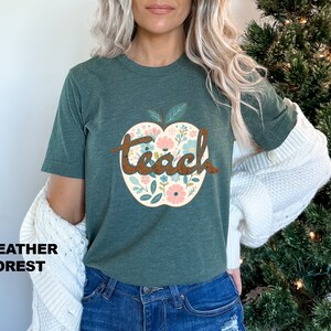Op de afbeelding: Een groen T-shirt met een bloemachtig appelontwerp en het woord "teach" in bruine tekst.