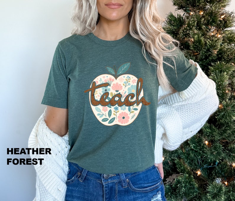 Op de afbeelding: Een groen T-shirt met een bloemachtig appelontwerp en het woord "teach" in bruine tekst.