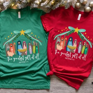 May include: Two t-shirts with a Christmas nativity scene design. The green shirt has the text "the greatest gift of all" and "Matthew 1:21". The red shirt has the text "the greatest gift of all" and "Matthew 1:21".