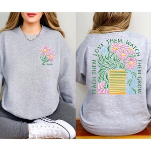 May include: A light gray sweatshirt featuring a floral design on the front and back. The front displays pink daisies and the text "Ms. Smith." The back showcases a vase of flowers with the text "Teach Them, Love Them, Watch Them Grow."