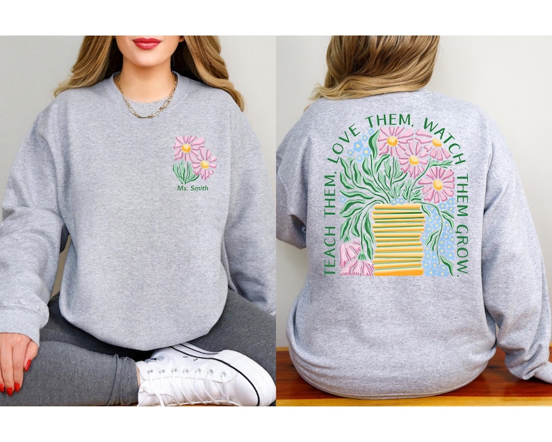 May include: A light gray sweatshirt featuring a floral design on the front and back. The front displays pink daisies and the text "Ms. Smith." The back showcases a vase of flowers with the text "Teach Them, Love Them, Watch Them Grow."