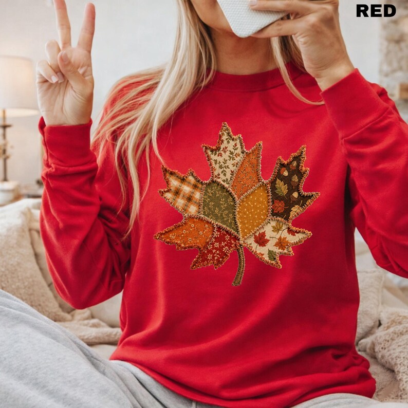 Pode incluir: Uma camisola vermelha de manga comprida com um grande gr&aacute;fico central de uma folha de &aacute;cer. A folha &eacute; composta por v&aacute;rios segmentos de tecido patchwork em cores de outono, incluindo laranja, castanho, verde e amarelo, com costuras vis&iacute;veis.