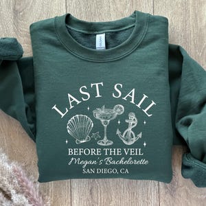 May include: A dark green sweatshirt with white text and graphics. The text reads "LAST SAIL BEFORE THE VEIL" with a seashell, cocktail, and anchor graphic. Below the text is "Megan's Bachelorette SAN DIEGO, CA".