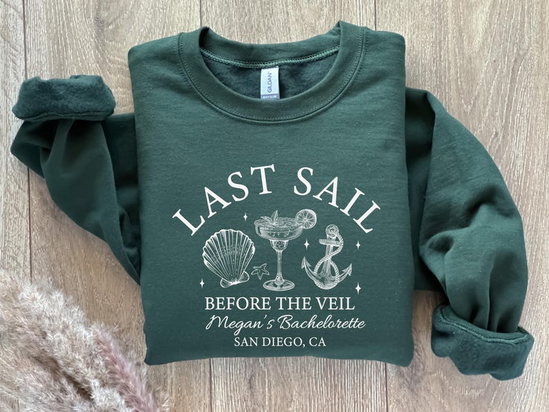 May include: A dark green sweatshirt with white text and graphics. The text reads "LAST SAIL BEFORE THE VEIL" with a seashell, cocktail, and anchor graphic. Below the text is "Megan's Bachelorette SAN DIEGO, CA".