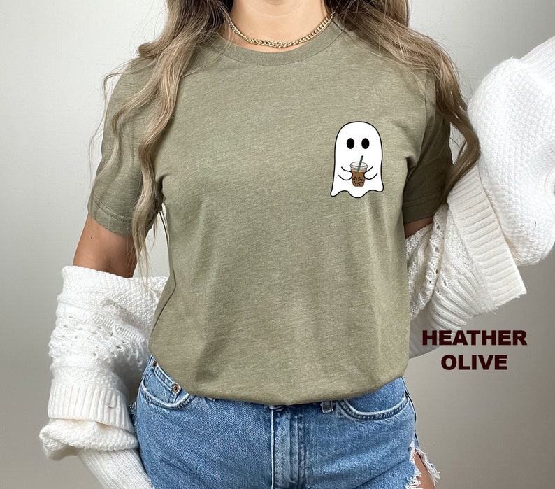 Puede incluir: Una camiseta de color oliva jaspeado con un gr&aacute;fico de un fantasma sosteniendo una taza de caf&eacute;. El texto "HEATHER OLIVE" est&aacute; impreso en la esquina inferior derecha de la imagen.